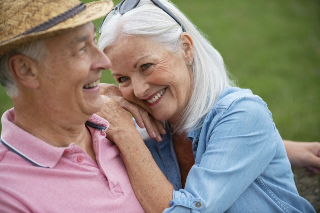 An older woman sitting, facing towards an old man. Her one arm over his, with her hands meeting on her shoulder, where she rests her head. She is smiling lovingly at the man who is smiling too while he looks in front of him.