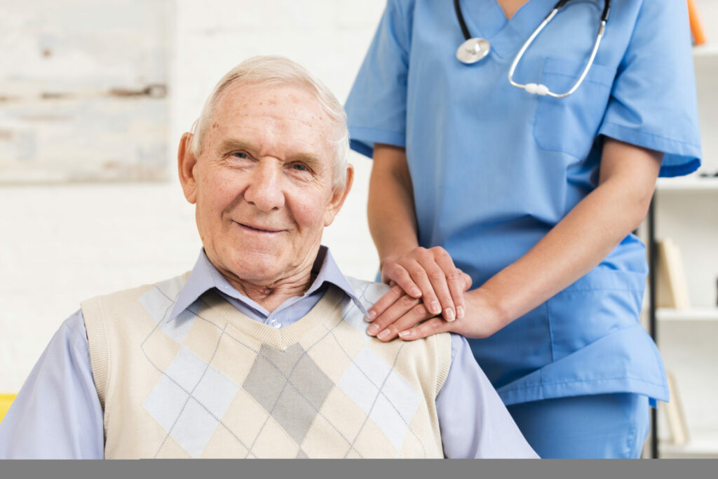 An old man sitting on a chair, with a nurse's hands placed on top of each other, resting on his shoulder.