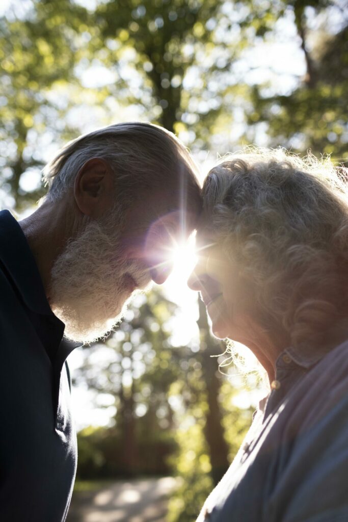 An elderly couple, standing facing each other, with their foreheads touching. the sun is shing through between their faces.