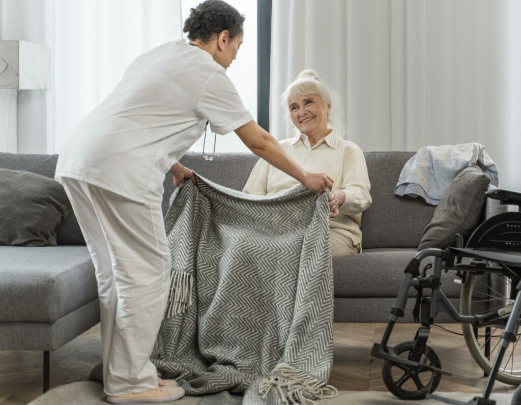 An elderly woman sitting on a couch while a nurse covers her with a blanket.