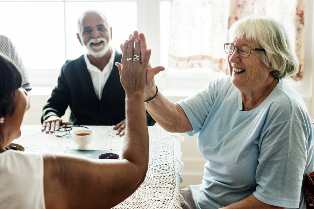 A group of old people sitting at a table, laughing. Two of the woman are giving each other a high five.